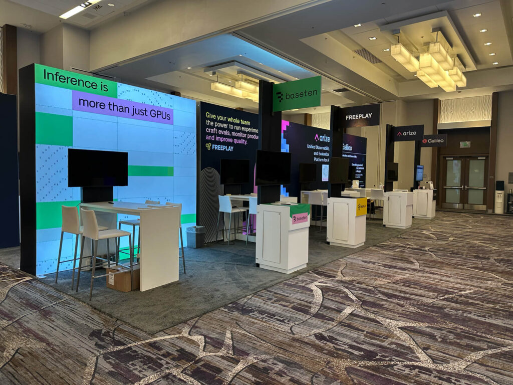 A line of sleek and modern exhibition booths at the ai engineer world's fair, featuring vibrant graphics with text like "inference is more than just gpus" and "basement," along with interactive screens and minimalist furniture.