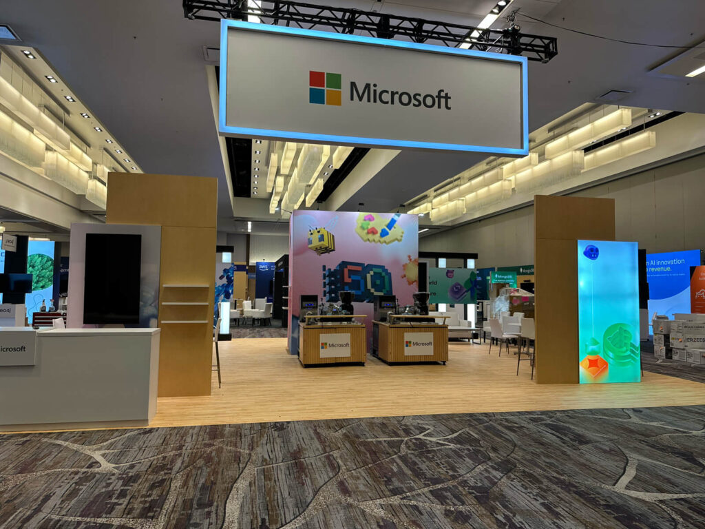 The microsoft exhibition booth at the ai engineer world's fair, featuring a large overhead illuminated sign, interactive display stations, colorful graphics, and a spacious layout with wooden flooring.