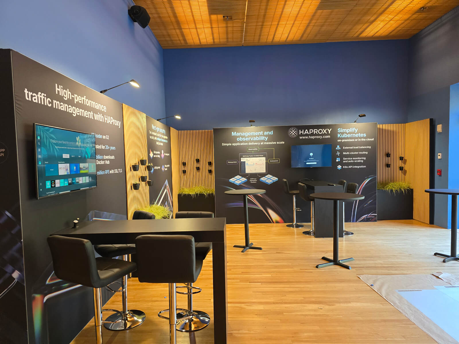 Detail shot of the haproxy event booth focusing on the "simplify kubernetes" section, showing text details, a monitor, and vertical slat wall elements.