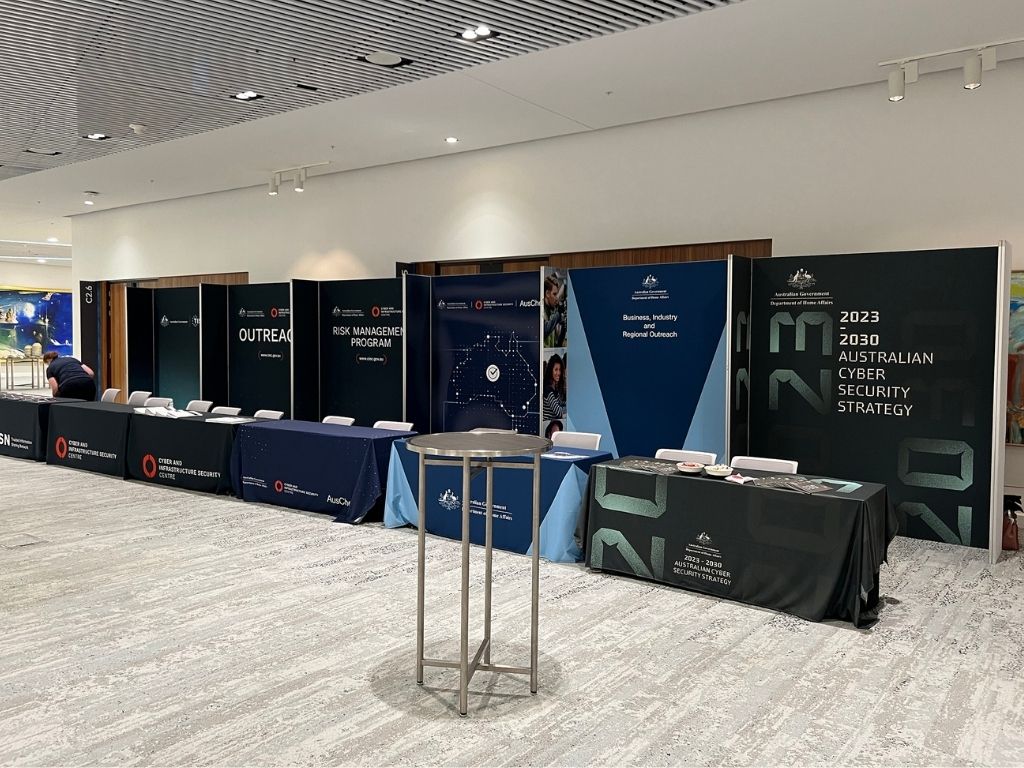 A modular exhibition booth setup for the australian government's cyber security strategy, featuring dark blue panels with logos behind draped tables in a brightly lit hall.