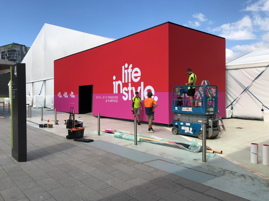 A man works on a building, with a large red sign prominently displayed in the background.