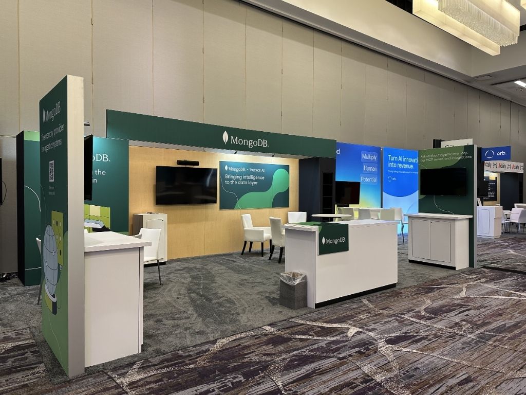 User-Inserted A modern, green and wood-toned mongodb exhibition stand featuring display screens, counters, and white seating.
