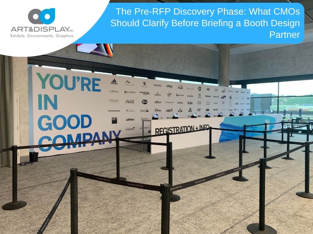 A trade show registration desk equipped with tablets, standing in front of a sponsor logo backdrop reading "You're in Good Company," with stanchions managing the queue.