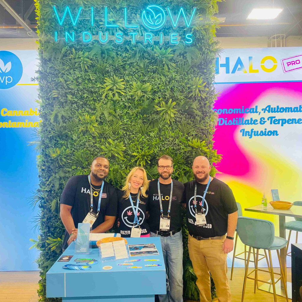 Willow industries trade show booth with a living plant wall, neon logo sign, and staff standing behind a blue product counter