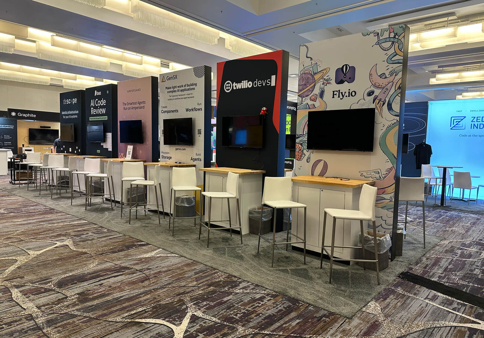 A row of modern, well-lit exhibition booths at the ai engineer world's fair, featuring prominent branding for "twilio dev/s" and "fly. Io," with high-top seating and interactive displays.